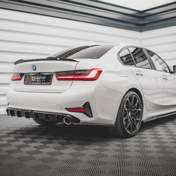 A white BMW sedan featuring sporty black wheels is parked, highlighting its dual exhaust and sharp rear design. The setting is a modern garage with textured walls and flooring. The text on the license plate reads "MAXTON DESIGN."