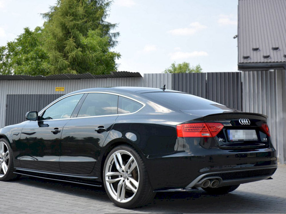 A black Audi car is parked outdoors on a paved surface with trees and a corrugated metal fence in the background. License plate details are blurred.
