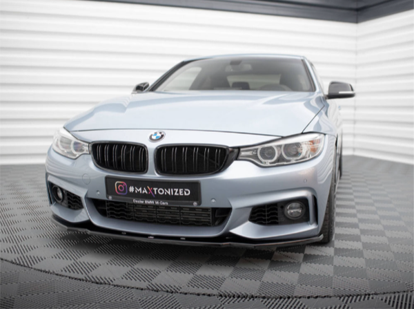 A silver BMW car is parked indoors, facing forward on a checkered floor. The license plate reads "#MAXTONIZED, Dealer BMW M Góra." The room has striped white and gray walls.