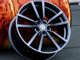 A black Volkswagen alloy wheel rests against a large orange cylinder with a wood-like texture on a gray surface.