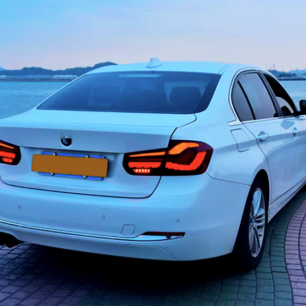A white sedan with illuminated taillights is parked on a patterned pavement beside a waterfront, with distant hills and a ship visible across a calm body of water.