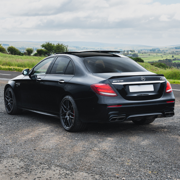 A black Mercedes-Benz sedan is parked on a rural road, surrounded by green fields under a cloudy sky. The text on the car reads "AMG" and "E52."