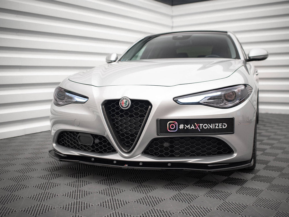 A silver car with sleek headlights and geometric grille stationary in a showroom-like setting with textured walls and floor. The text on a plate reads: "@MAXTONIZED".