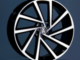 A black and silver alloy wheel, featuring the Volkswagen logo at its center, is displayed upright against a neutral gray background on a reflective surface.