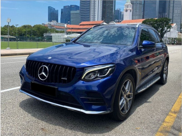 A blue Mercedes-Benz SUV is parked on a city street. Skyscrapers and a clock tower appear in the background, with greenery and a clear sky enhancing the urban setting.