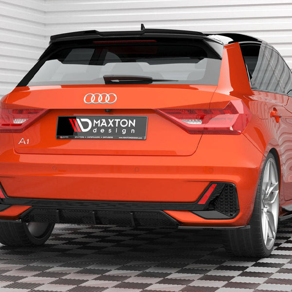 A bright orange Audi A1 is parked in a garage with white paneled walls and checkered flooring. The license plate reads "MAXTON design." The car features sleek, sporty lines.
