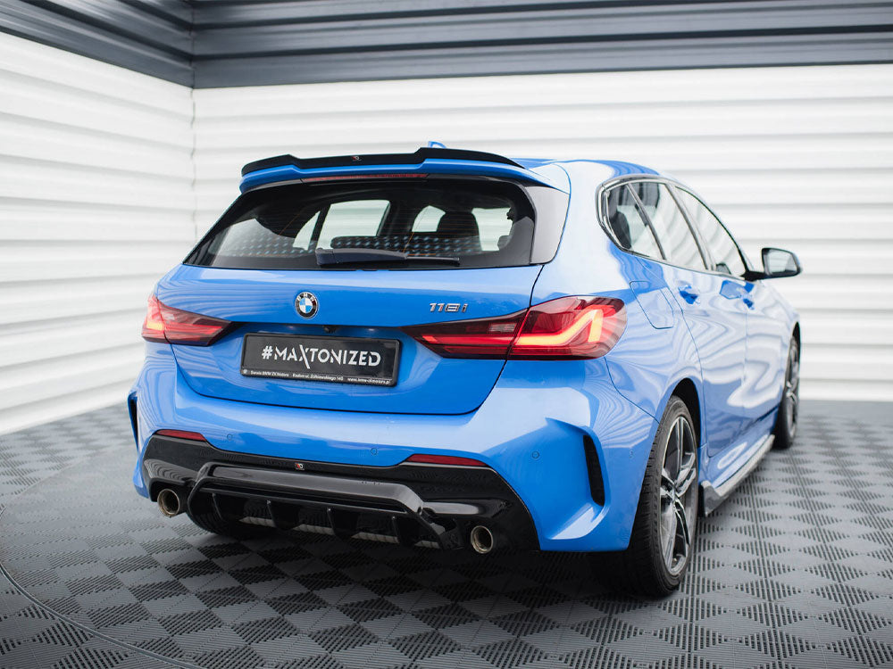 A blue BMW 116i parked, rear view, with illuminated taillights, in a showroom with a geometric-patterned floor and striped walls. License plate reads "#MAXTONIZED".