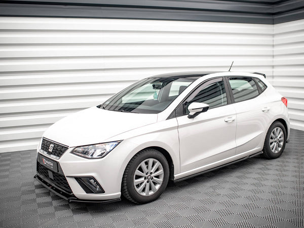 A white hatchback car is parked in a modern, well-lit showroom with patterned flooring and walls.