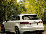 A white Audi S3 car is parked on a leaf-strewn path, surrounded by lush green and autumn-hued trees, creating a serene outdoor setting.