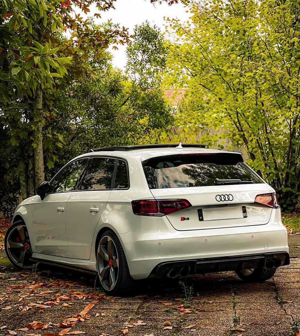 A white Audi S3 car is parked on a leaf-strewn path, surrounded by lush green and autumn-hued trees, creating a serene outdoor setting.