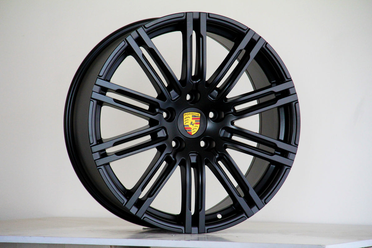 A black alloy wheel, featuring a multi-spoke design and the Porsche emblem at the center, stands against a plain background, resting on a white surface.