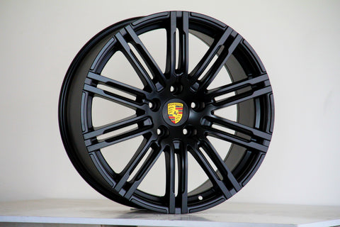 A black alloy wheel, featuring a multi-spoke design and the Porsche emblem at the center, stands against a plain background, resting on a white surface.