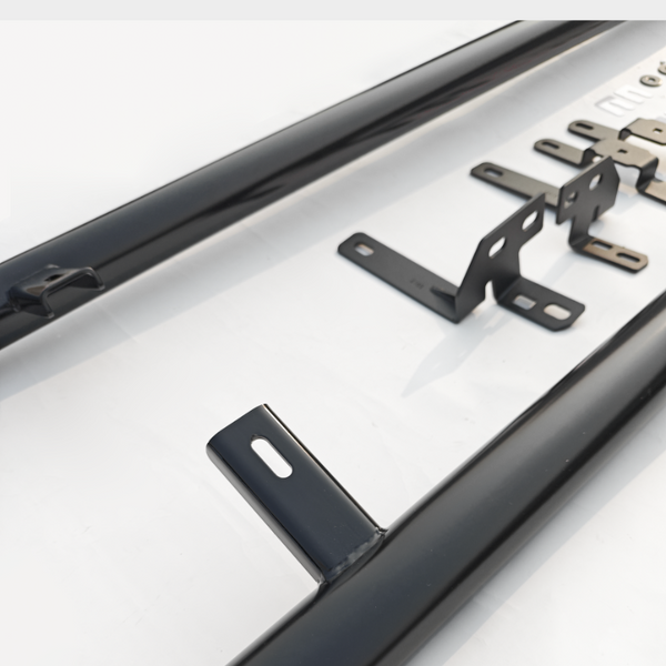 Black metal bars with mounting brackets are laid out on a white surface. The brackets have multiple holes and are positioned beside small washers and screws, suggesting assembly.