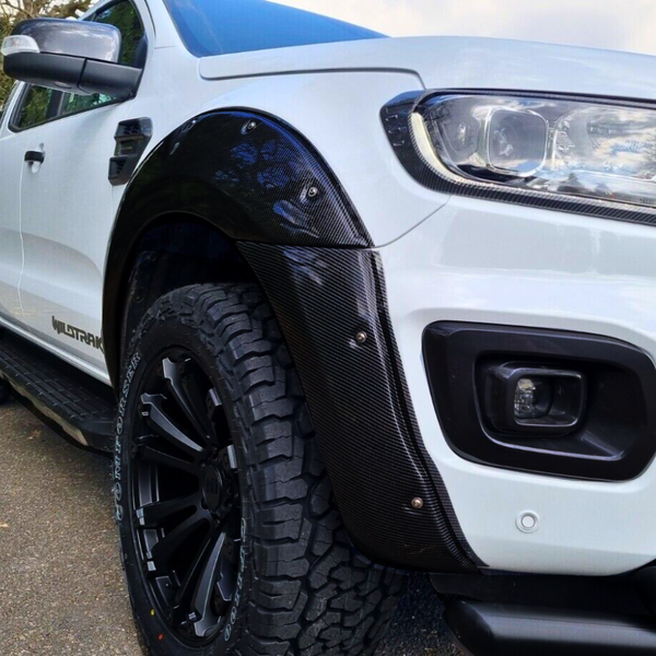 A white pickup truck features a black carbon fiber wheel arch, chunky off-road tires, and "WILDTRAK" text on the side. It's parked on a street with trees visible in the background.