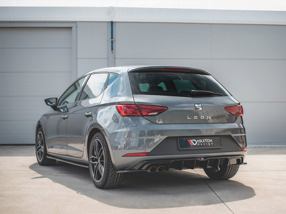 A grey SEAT Leon car is parked on a concrete surface near a white building. The rear features a Maxton Design diffuser and visible dual exhausts.