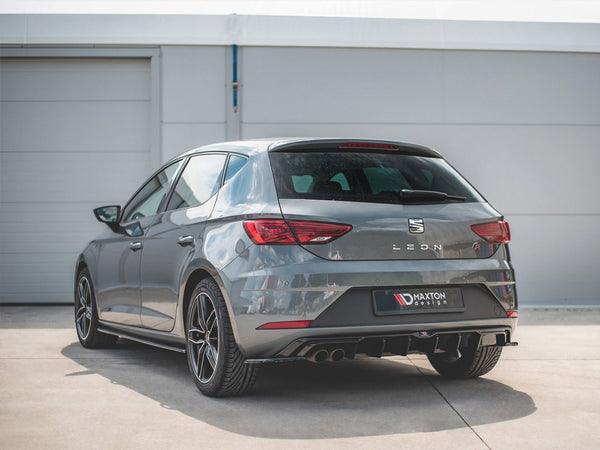 A grey SEAT Leon car is parked on a concrete surface near a white building. The rear features a Maxton Design diffuser and visible dual exhausts.