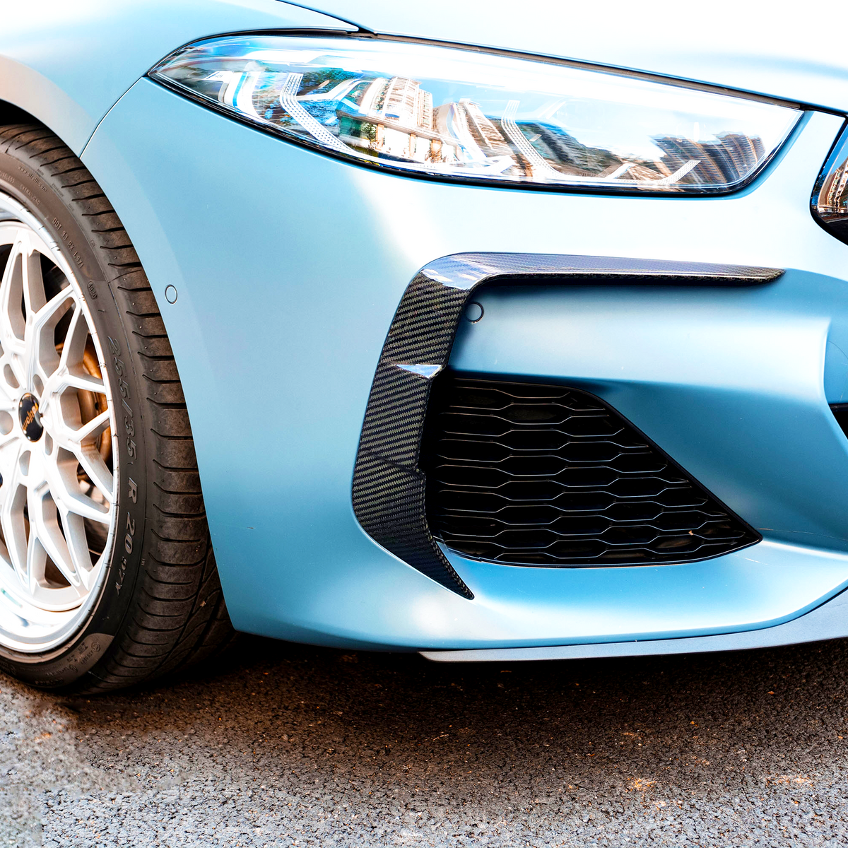 A close-up of a blue car's front, featuring a sleek headlight, sporty grille, and detailed alloy wheel. The vehicle is parked on a paved surface.