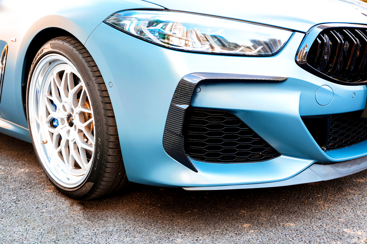 A close-up of a blue car's front, featuring a sleek headlight, sporty grille, and detailed alloy wheel. The vehicle is parked on a paved surface.