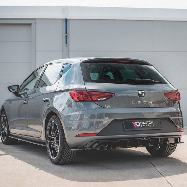 A gray hatchback car is parked on a concrete surface in front of a garage. The rear displays "LEON" and "MAXTON design" text, with visible dual exhausts and stylish rims.