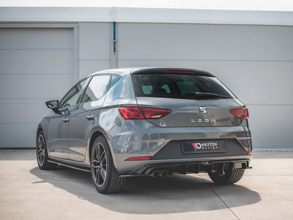 A gray hatchback car is parked on a concrete surface in front of a garage. The rear displays "LEON" and "MAXTON design" text, with visible dual exhausts and stylish rims.