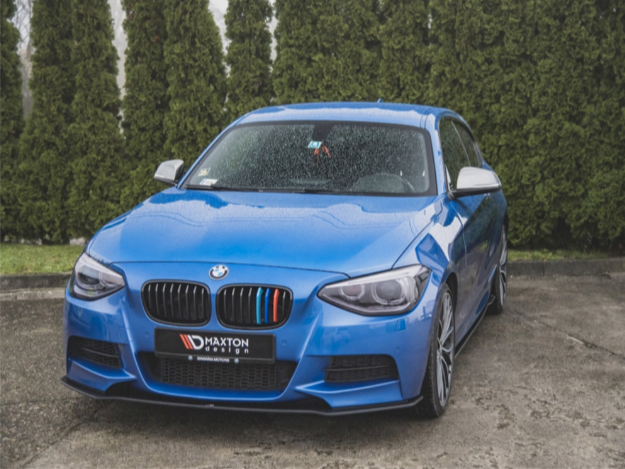 A blue BMW car parked on wet pavement, displaying a front license plate reading "MAXTON DESIGN," with a backdrop of tall green bushes.