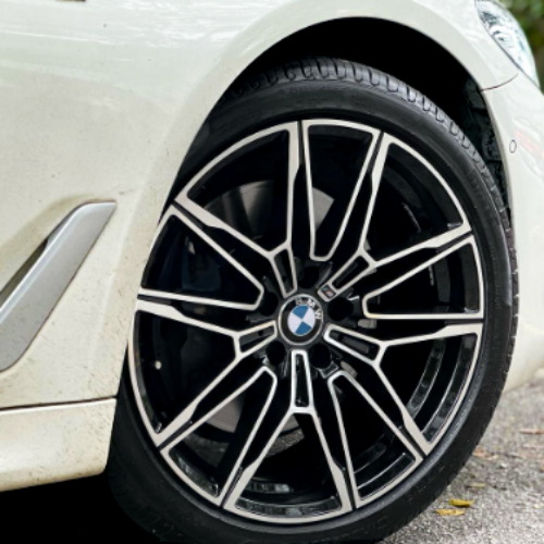 A car wheel with a BMW logo is stationary on a black tire, featuring intricate black and silver alloy rim designs. It's positioned on a paved road, part of a white vehicle.