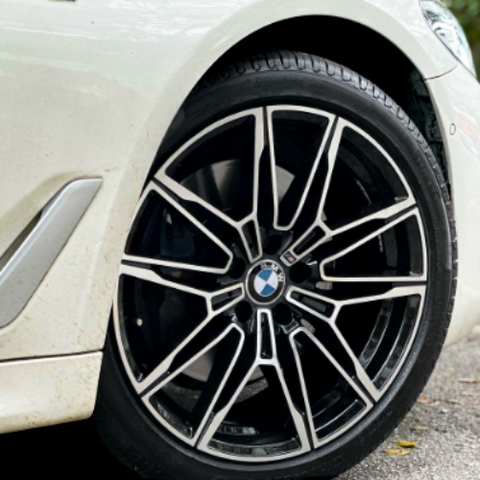 A car wheel with a BMW logo is stationary on a black tire, featuring intricate black and silver alloy rim designs. It's positioned on a paved road, part of a white vehicle.