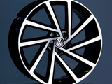A black and white alloy wheel, featuring a Volkswagen logo in the center, stands on a round podium against a gray background.