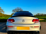 A silver Mercedes-Benz car is parked on a paved road, with its rear facing open fields and clear blue sky. The logo is visible on the trunk.