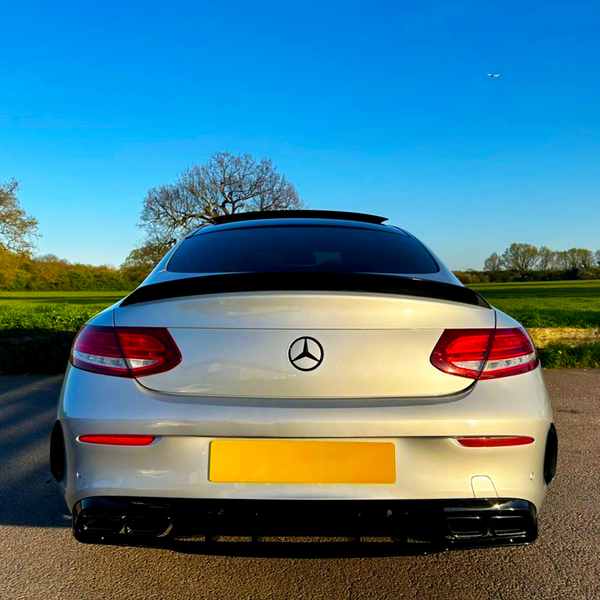 A silver Mercedes-Benz car is parked on a paved road, with its rear facing open fields and clear blue sky. The logo is visible on the trunk.