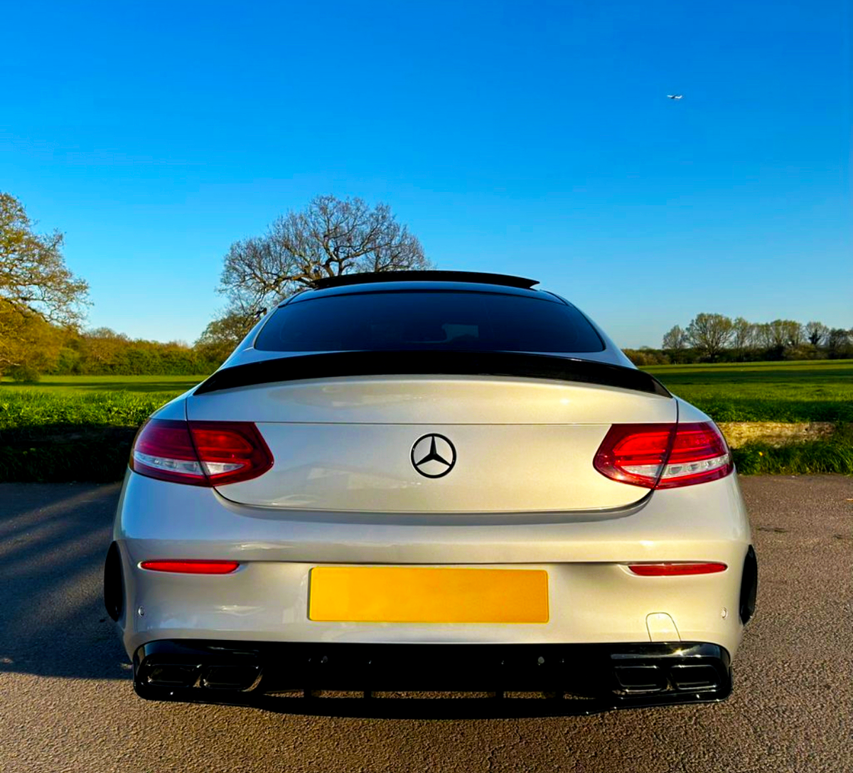 A silver Mercedes-Benz car is parked on a paved road, with its rear facing open fields and clear blue sky. The logo is visible on the trunk.