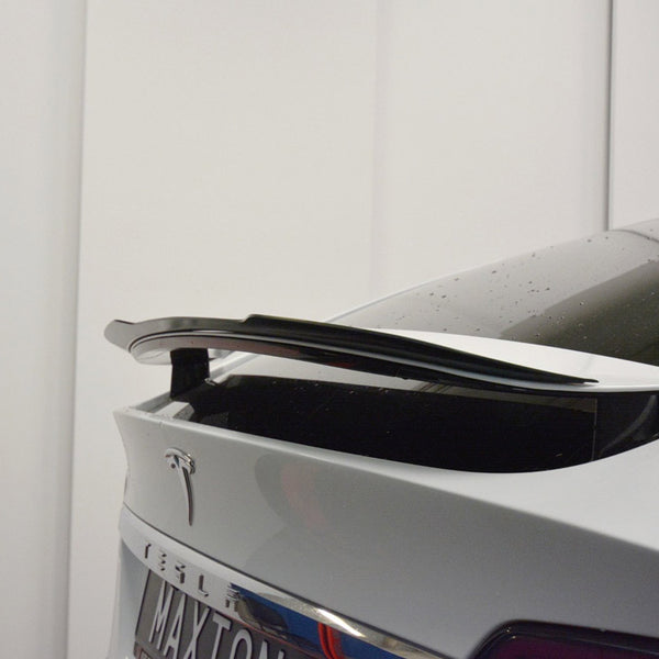 A car's rear end features a black spoiler on a white polished surface, with "MA-XTON" visible on the license plate. The background is a plain white wall.