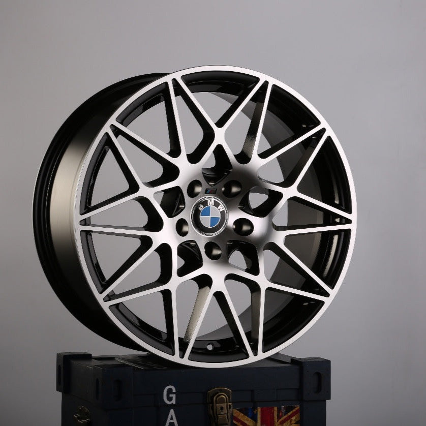 A BMW car rim with an intricate, geometric design stands upright on a dark blue box emblazoned with the letters "GA" and a partial Union Jack in a plain gray environment.