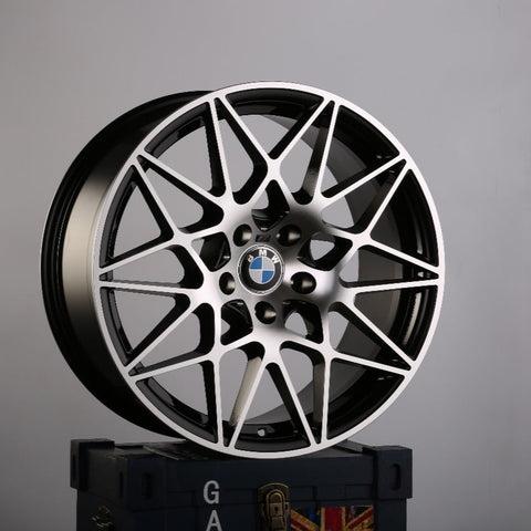 A BMW car rim with an intricate, geometric design stands upright on a dark blue box emblazoned with the letters "GA" and a partial Union Jack in a plain gray environment.