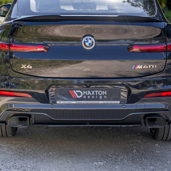 A black BMW X4 M40i is parked on a gravel surface. The license plate reads "MAXTON design." The car's rear includes dual exhausts and prominent taillights.