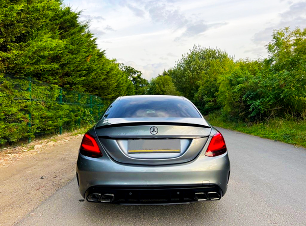 A silver Mercedes-Benz car is parked on a road, surrounded by lush green trees under a partly cloudy sky.