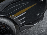 A car tire with a black alloy wheel is parked on asphalt. The vehicle features carbon fiber aerodynamic fins and a glossy black surface, reflecting a yellow line. Text: 