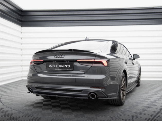 A sleek, dark gray Audi car is parked in a showroom with checkered flooring. The license plate reads "#MAJTONIZED." The room has striped gray and white walls.