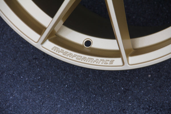 A gold car rim with the text "MPERFORMANCE" engraved, shown close-up on a textured black surface.