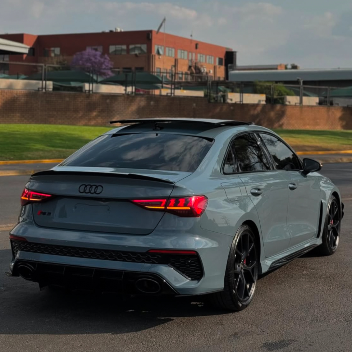 A gray Audi RS3 is parked on a paved surface. Its rear lights are illuminated. The background features a red brick building, trees, and a grassy area under a cloudy sky.