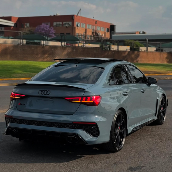 A gray Audi RS3 is parked on a paved surface. Its rear lights are illuminated. The background features a red brick building, trees, and a grassy area under a cloudy sky.