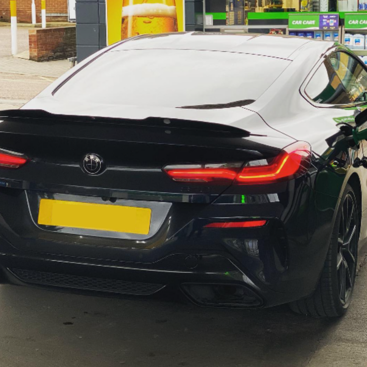 A black sports car is refueling at a gas station. The car's rear lights are illuminated, and shelves with automotive products are visible in the background.