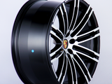 A glossy black and silver alloy wheel is angled to show its intricate spoke design, featuring a central emblem with the Porsche logo. The background is plain white.