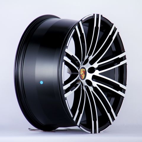 A glossy black and silver alloy wheel is angled to show its intricate spoke design, featuring a central emblem with the Porsche logo. The background is plain white.