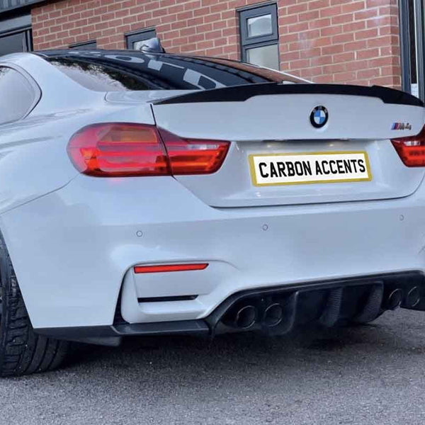 A white BMW car is parked, displaying its black wheels and dual exhausts. It has a license plate reading "CARBON ACCENTS." The setting features a brick building and a glass door.