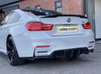 A white BMW car is parked, displaying its black wheels and dual exhausts. It has a license plate reading "CARBON ACCENTS." The setting features a brick building and a glass door.
