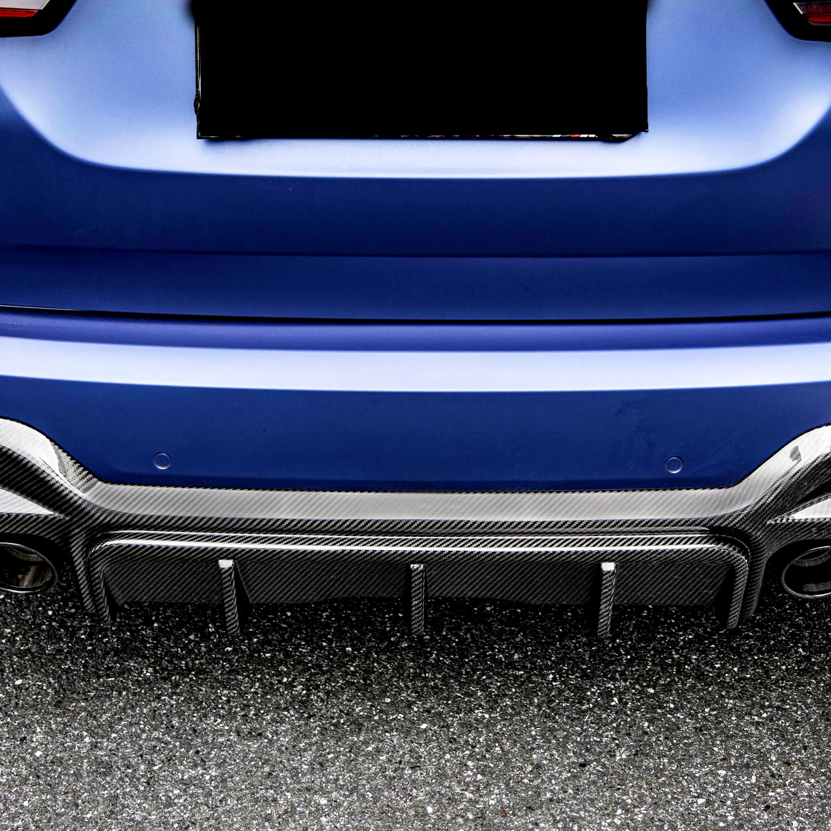 A blue car's rear with a carbon fiber diffuser and four exhaust pipes, parked on an asphalt surface. The license plate area is obscured.
