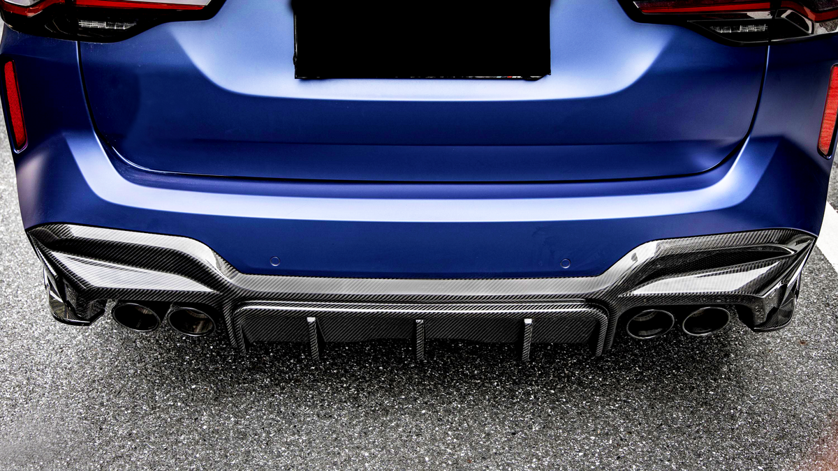 A blue car's rear with a carbon fiber diffuser and four exhaust pipes, parked on an asphalt surface. The license plate area is obscured.
