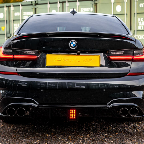 A black BMW with visible exhaust pipes, parked on gravel, is positioned in front of green storage containers. The taillights are illuminated, and the license plate is intentionally blurred out.