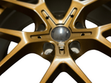 A bronze alloy wheel with a multi-spoke design lies against a white background. The central hub area is visible, surrounded by intricate metal detailing.
